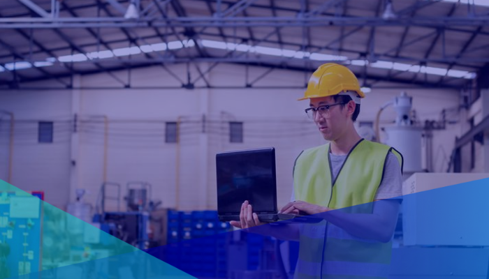 A man in a hard hat is using a laptop in a factory to bridge knowledge gaps between automation and aging workforces.