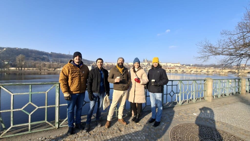 Five people in winter clothing stand smiling by a river with a scenic city and bridge in the background under a clear blue sky.