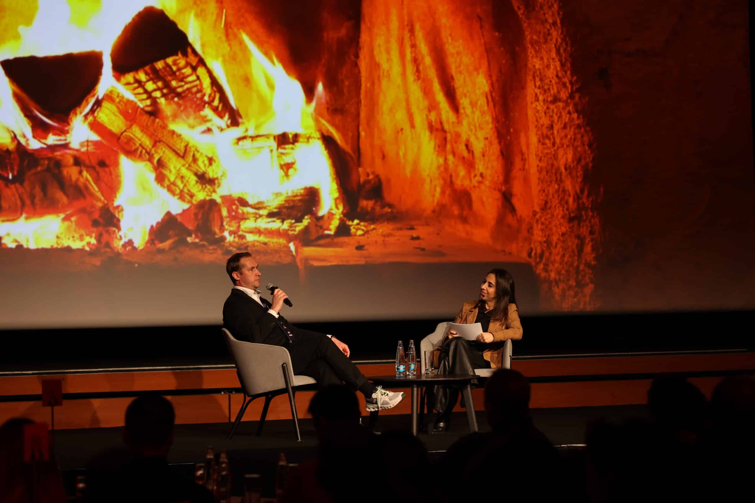 Two people sit on stage in armchairs having a conversation; one holds a microphone, the other has papers. A large image of a burning fireplace is displayed behind them, creating a warm, cozy atmosphere. Bottles and glasses sit on the table.