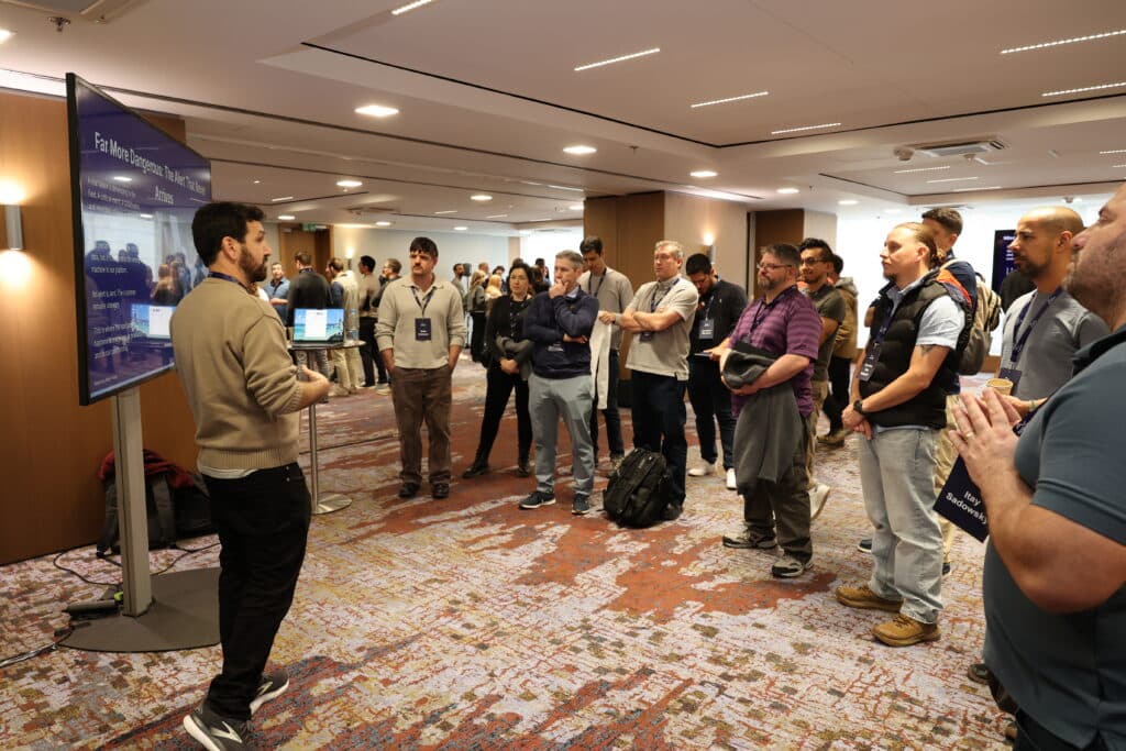 A man stands next to a large screen and speaks to a group of people gathered around him in a conference room, suggesting a presentation or workshop setting.
