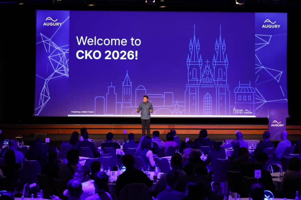 A speaker stands on stage in front of a large screen that reads “Welcome to CKO 2026!” with the Augury logo, addressing a seated audience in a dimly lit conference room.