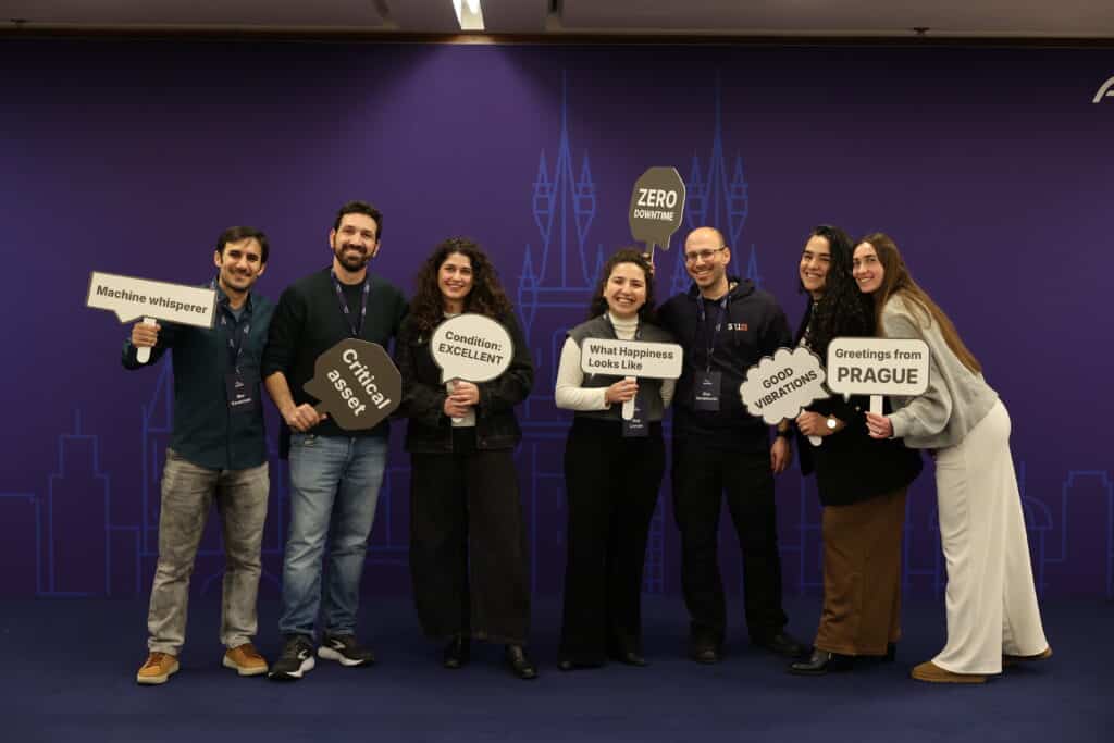 Seven people stand smiling in front of a purple backdrop, holding signs with phrases like “Machine whisperer,” “Critical sense,” “What Happiness Looks Like,” and “Greetings from PRAGUE.”.