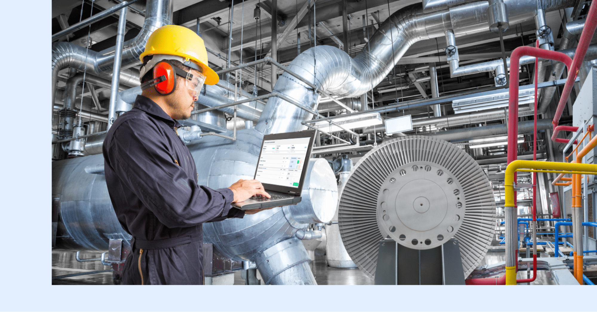 A worker in safety gear uses a laptop while standing in an industrial facility filled with large metal pipes and machinery.