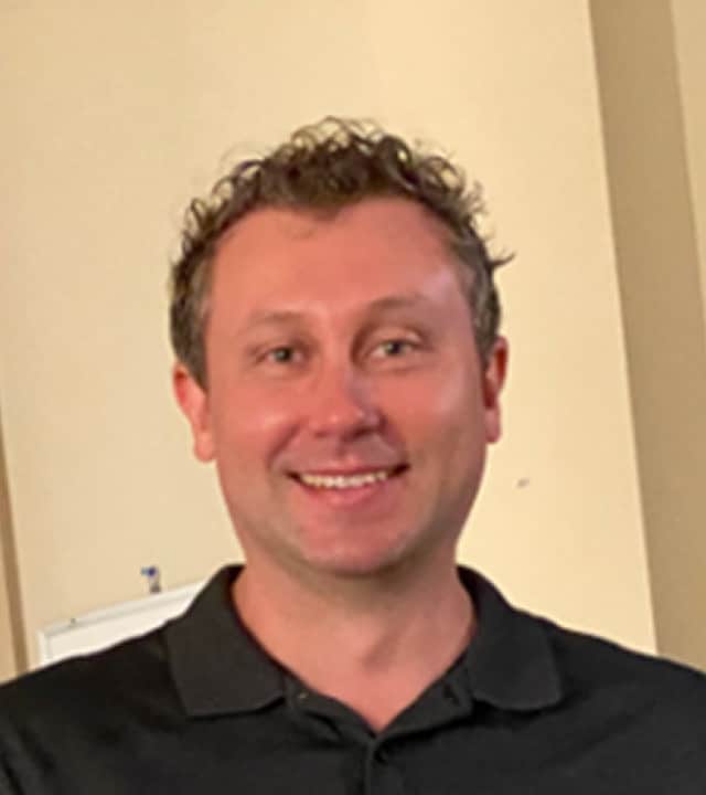 A man with short, curly light brown hair and blue eyes is smiling at the camera. He is wearing a black collared shirt and is standing indoors against a beige wall.