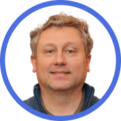 Portrait of a middle-aged man with light brown curly hair, light skin, and a slight smile, wearing a dark jacket. The image is framed by a blue circular border on a white background.