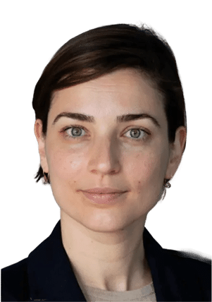 A woman with short brown hair, light eyes, and a neutral expression is facing the camera. She is wearing a dark blazer over a light top and small earrings, with a plain white background.