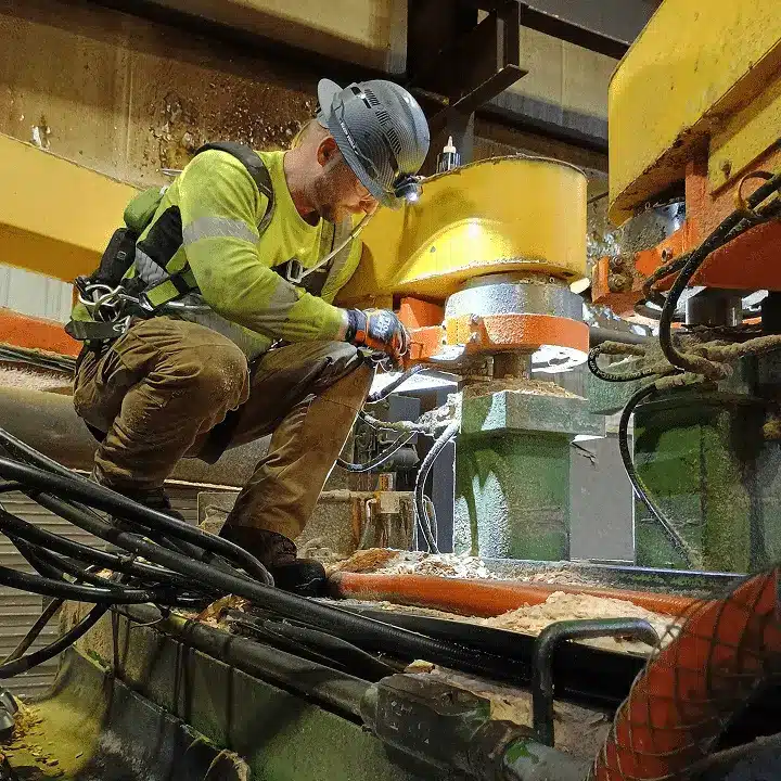 A construction worker in safety gear kneels on machinery, using tools to work on industrial equipment inside a factory or plant. Cables and large metal structures surround him.