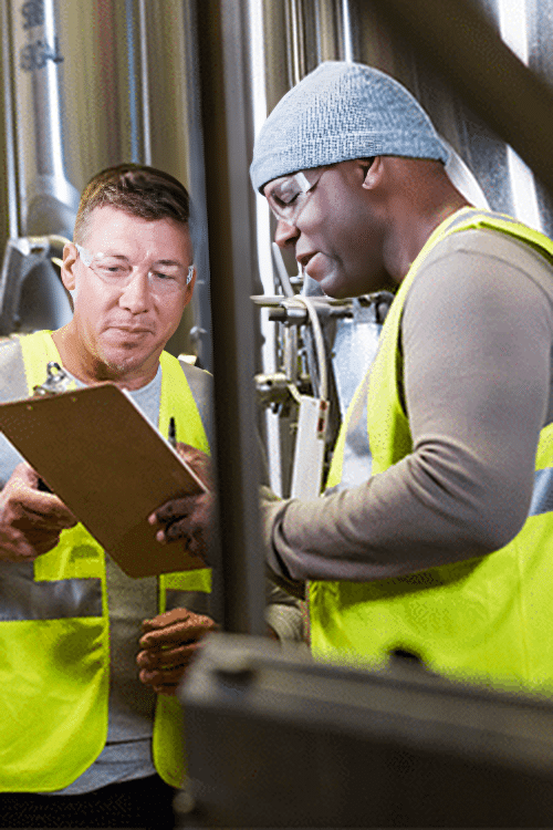 Two men in safety vests and casual clothing examine a clipboard together in an industrial setting, possibly a factory or warehouse, where Augury's AI empowers their decision-making process.