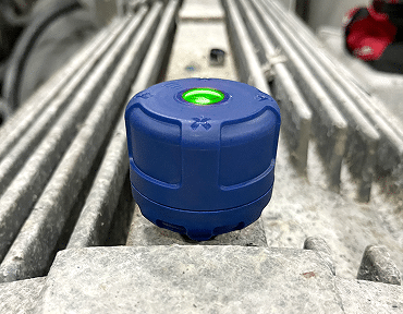 A blue, round electronic device with a green indicator light sits on a dusty, ribbed metal surface in an industrial setting.