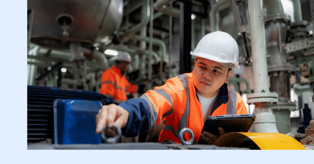 Worker in an orange safety jacket and white helmet inspects industrial machinery while holding a tablet; another worker is visible in the background amid pipes and equipment.