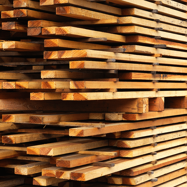 A close-up view of neatly stacked wooden planks, showing their grain patterns and edges. The boards are arranged horizontally in rows, highlighting the natural color and texture of the wood.