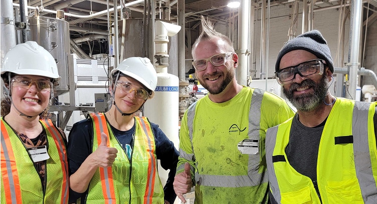 Four people wearing safety vests, hard hats, and protective glasses pose and smile inside an industrial facility. Two give thumbs-up gestures. Machinery and large pipes are visible in the background.
