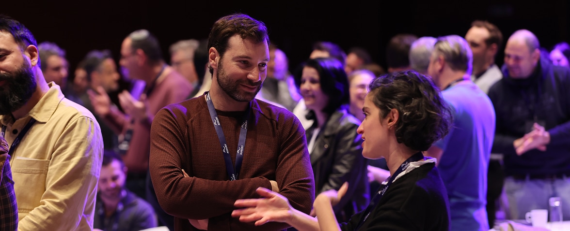 A man with a lanyard listens with arms crossed as a woman gestures while speaking to him, surrounded by a diverse crowd engaged in conversation at a lively indoor event.