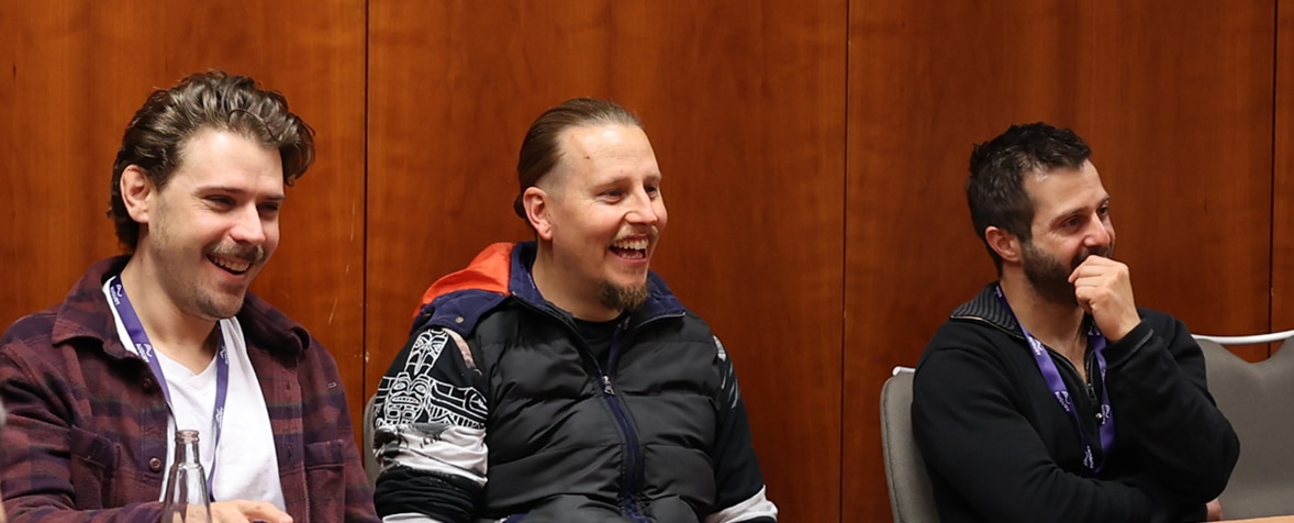 Three men sit in chairs indoors, smiling and laughing together. They wear casual clothes and lanyards, and are seated in front of a wooden wall.