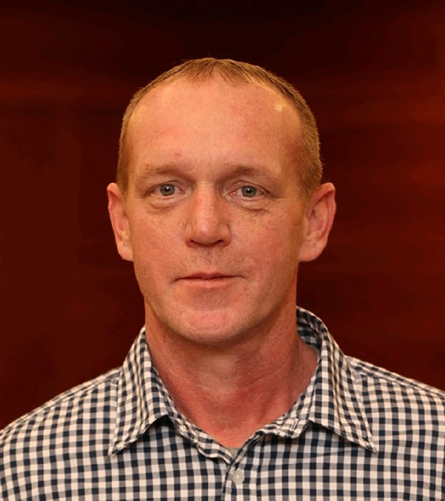 A man with short light brown hair is wearing a black-and-white checkered shirt and is looking at the camera, with a dark reddish-brown background behind him.