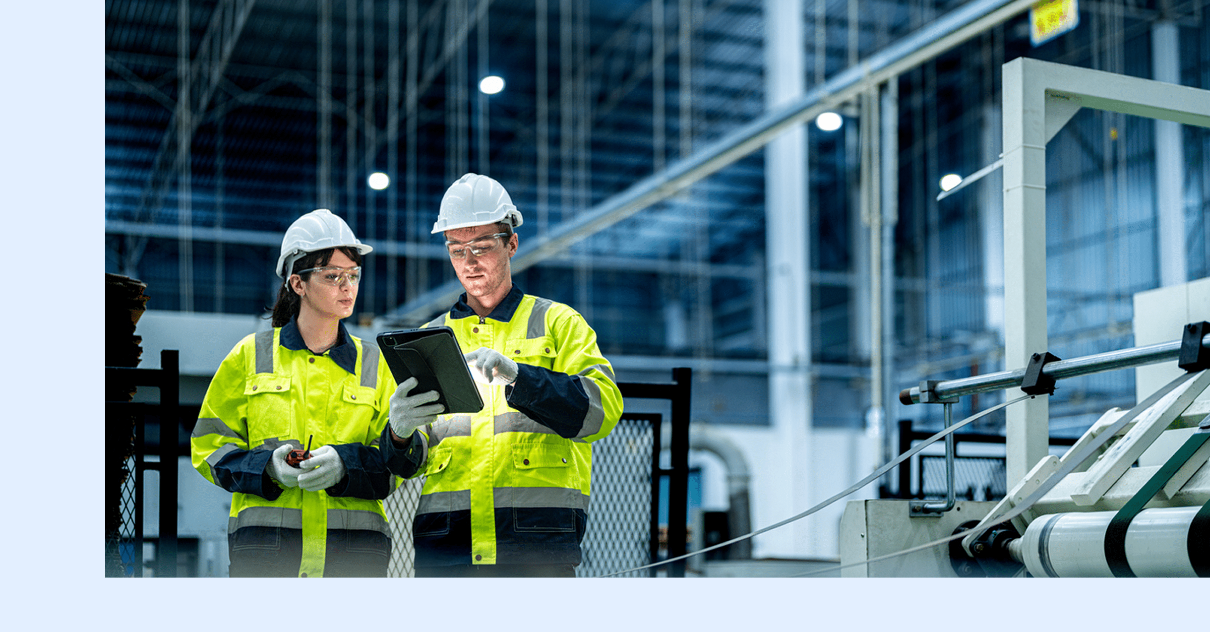 Two workers in safety gear and helmets review information on a tablet inside a modern industrial facility, surrounded by machinery and metal structures.