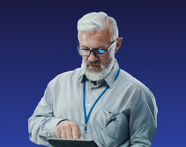 An older man with white hair and beard, wearing glasses and a gray shirt with a lanyard, is using a tablet against a plain blue background.