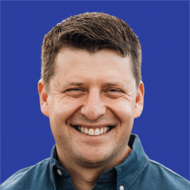 A man with short brown hair smiles broadly, wearing a dark blue collared shirt, against a solid blue background.