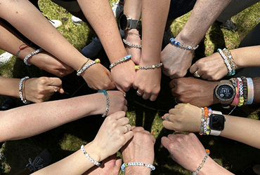 A group of people stand in a circle, extending their arms inward to display matching beaded bracelets on their wrists. The image is taken outdoors on grass and shows diverse hands and wrists.