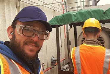 Two workers wearing safety gear, including hard hats, safety glasses, and orange vests, are outside near industrial equipment. One faces the camera and smiles; the other has their back turned while working.