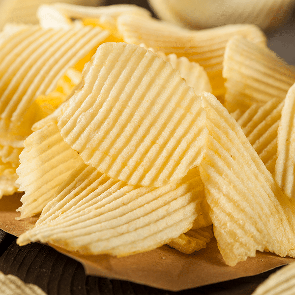 A close-up of a pile of ridged, golden potato chips on a brown surface, showing their crisp texture and wavy edges.