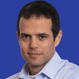 A man with short dark hair, wearing a light blue collared shirt, looks directly at the camera with a neutral expression. The background is solid blue.