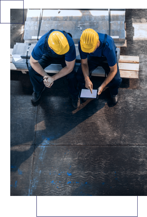 Two workers wearing yellow hard hats and blue uniforms sit on a stack of materials, with one reviewing something on a tablet to make smarter decisions.
