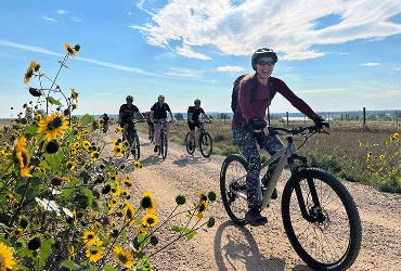 Four people ride bikes on a dirt trail beside blooming sunflowers under a bright blue sky. The lead cyclist smiles, wearing a helmet and sunglasses, with fields and a fence in the background.
