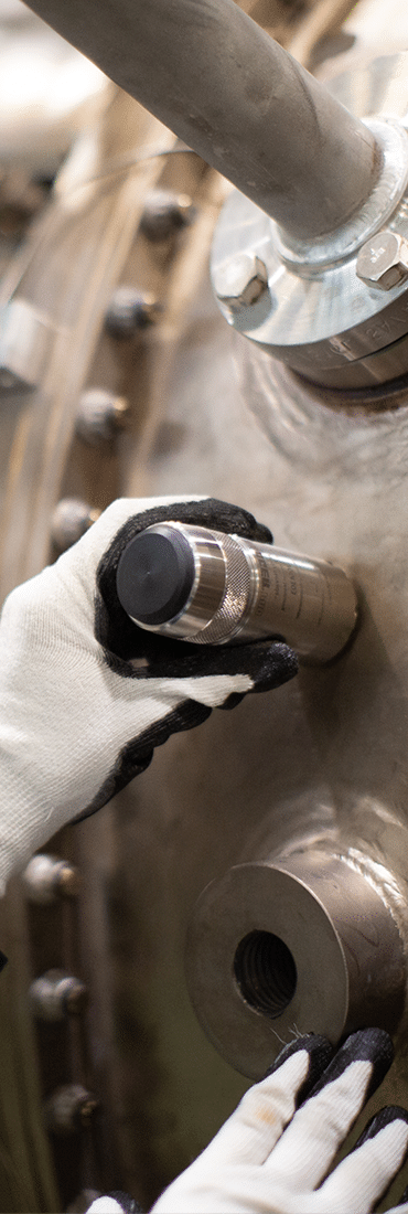 Gloved hands are attaching or inspecting a metallic cylindrical component on a large industrial machine with bolts and pipes.