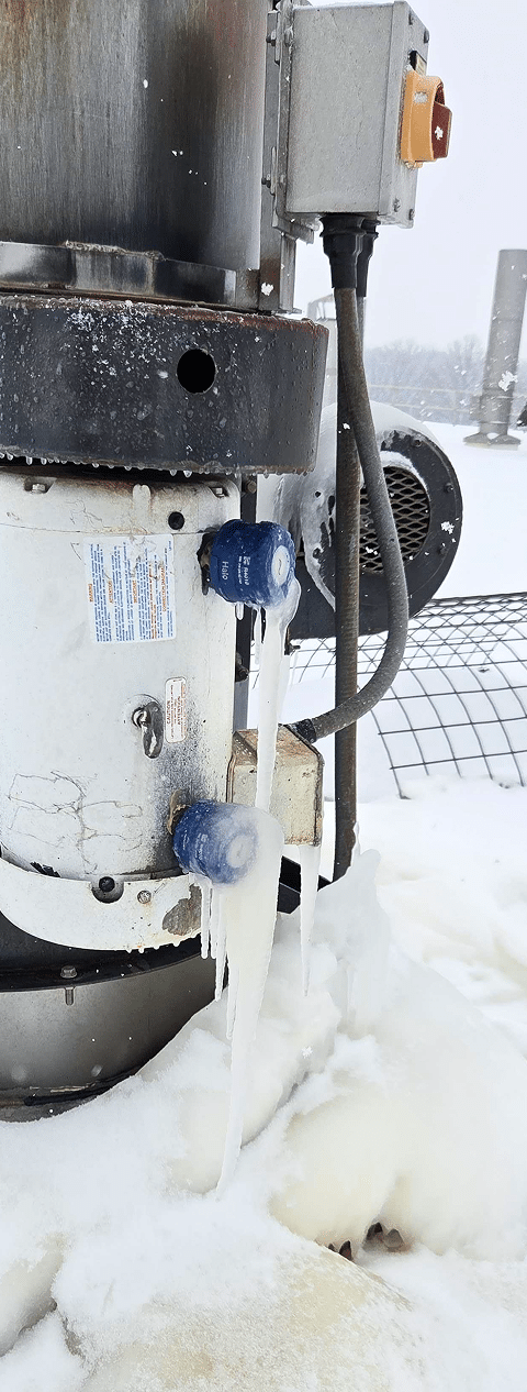 A close-up of industrial equipment outdoors in winter, with long icicles hanging from blue valve handles. Snow and ice cover parts of the machine and ground, with a wire fence and snowy landscape in the background.