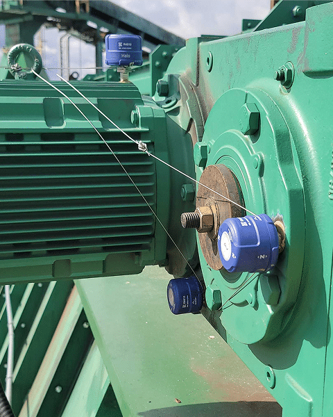 Close-up of industrial machinery with blue sensor devices attached to a green metal motor and gearbox, connected by thin wires. The background includes more machinery and a partly cloudy sky.