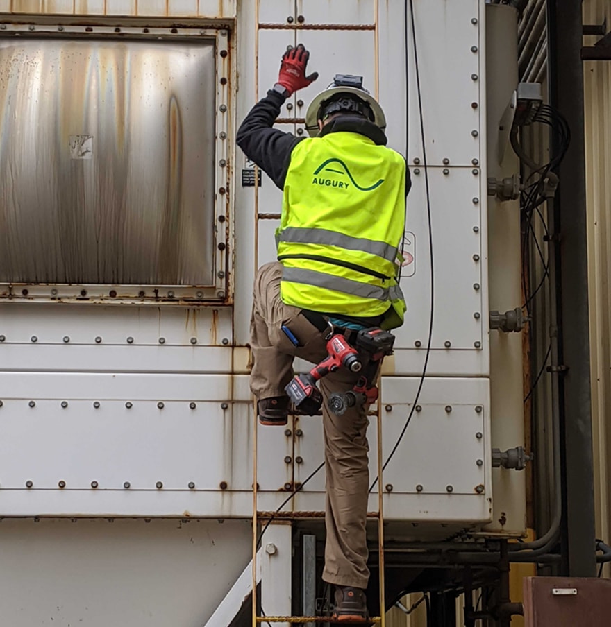 A worker wearing a neon yellow safety vest and helmet climbs a metal ladder attached to industrial equipment, holding a power tool and raising one hand to touch the structure.