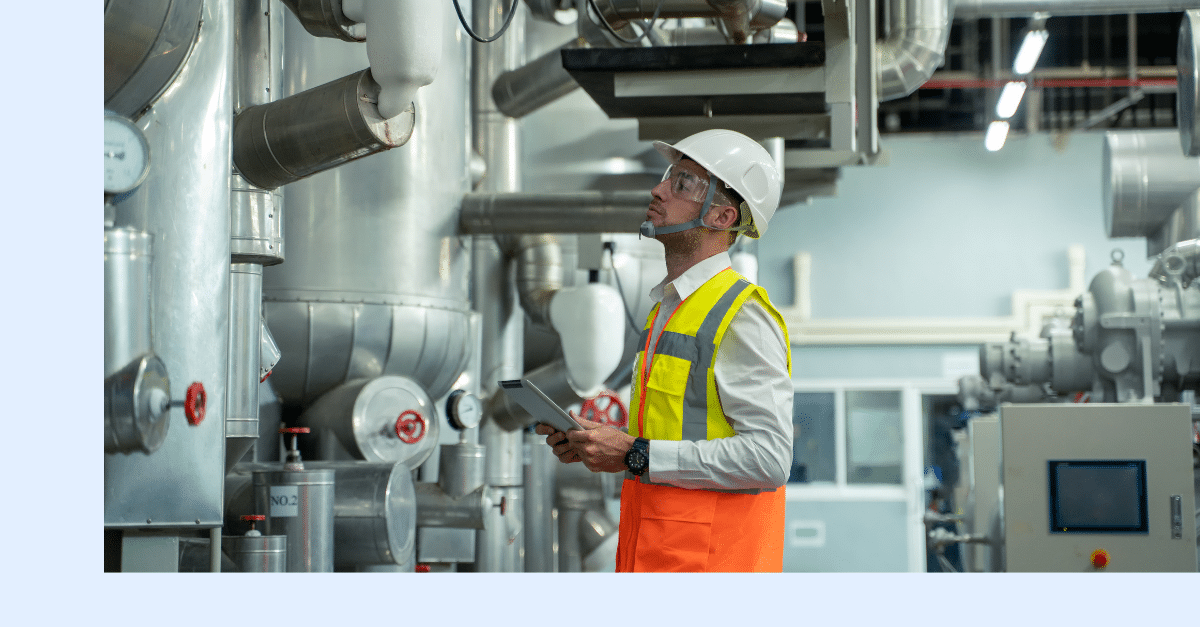 A male engineer in a hard hat and high-visibility vest inspects industrial equipment in a factory, holding a tablet and looking up at large pipes and machinery.