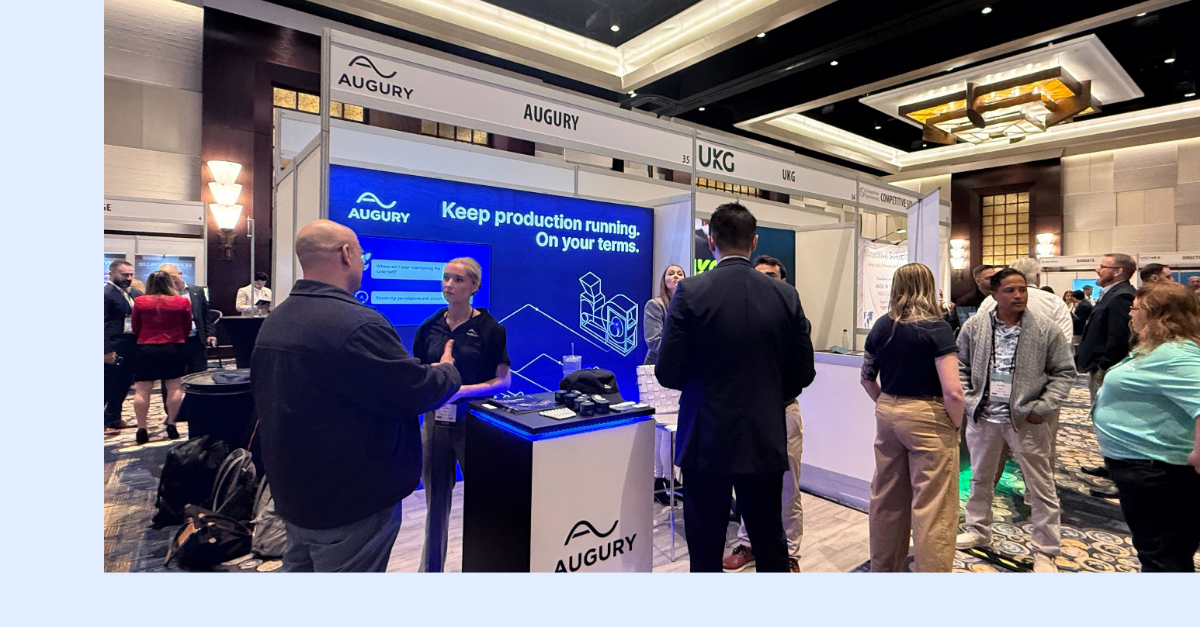 People interact at the Augury booth during a business expo. A large digital display reads “Keep production running. On your terms.” Several attendees are engaged in conversation, and exhibits are visible in the background.