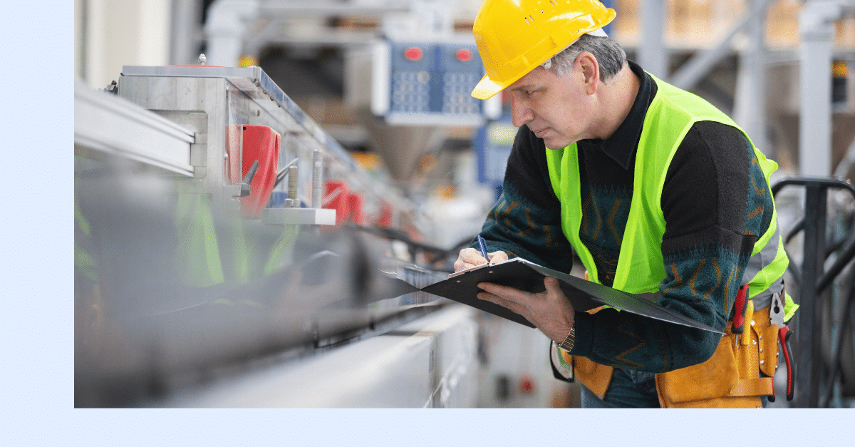 A man wearing a yellow hard hat, safety vest, and tool belt writes on a clipboard while inspecting machinery in an industrial setting.