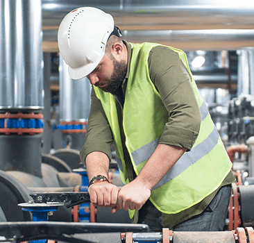 A man wearing a white hard hat and a yellow reflective safety vest is using a large wrench to adjust industrial pipes in a facility.