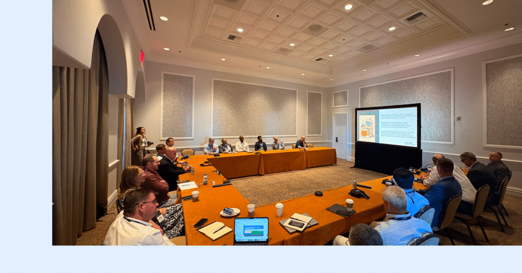 A group of people are seated around a large U-shaped table in a conference room, watching a presentation displayed on a large screen at the front of the room. Laptops, coffee cups, and papers are on the tables.