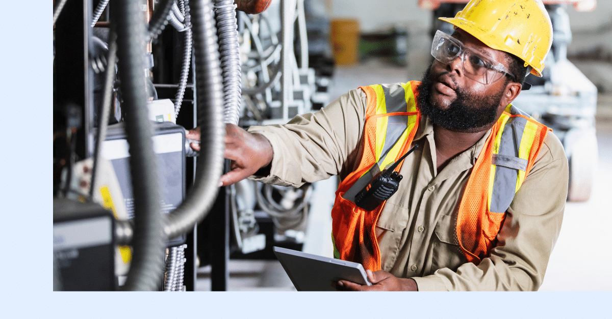 A worker wearing a yellow hard hat, safety glasses, and an orange safety vest inspects electrical equipment while holding a tablet in an industrial setting.
