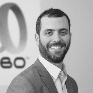 Black and white photo of a man with a beard, wearing a suit, smiling at the camera. A partial logo is visible in the background, hinting at his commitment to Machine Health.