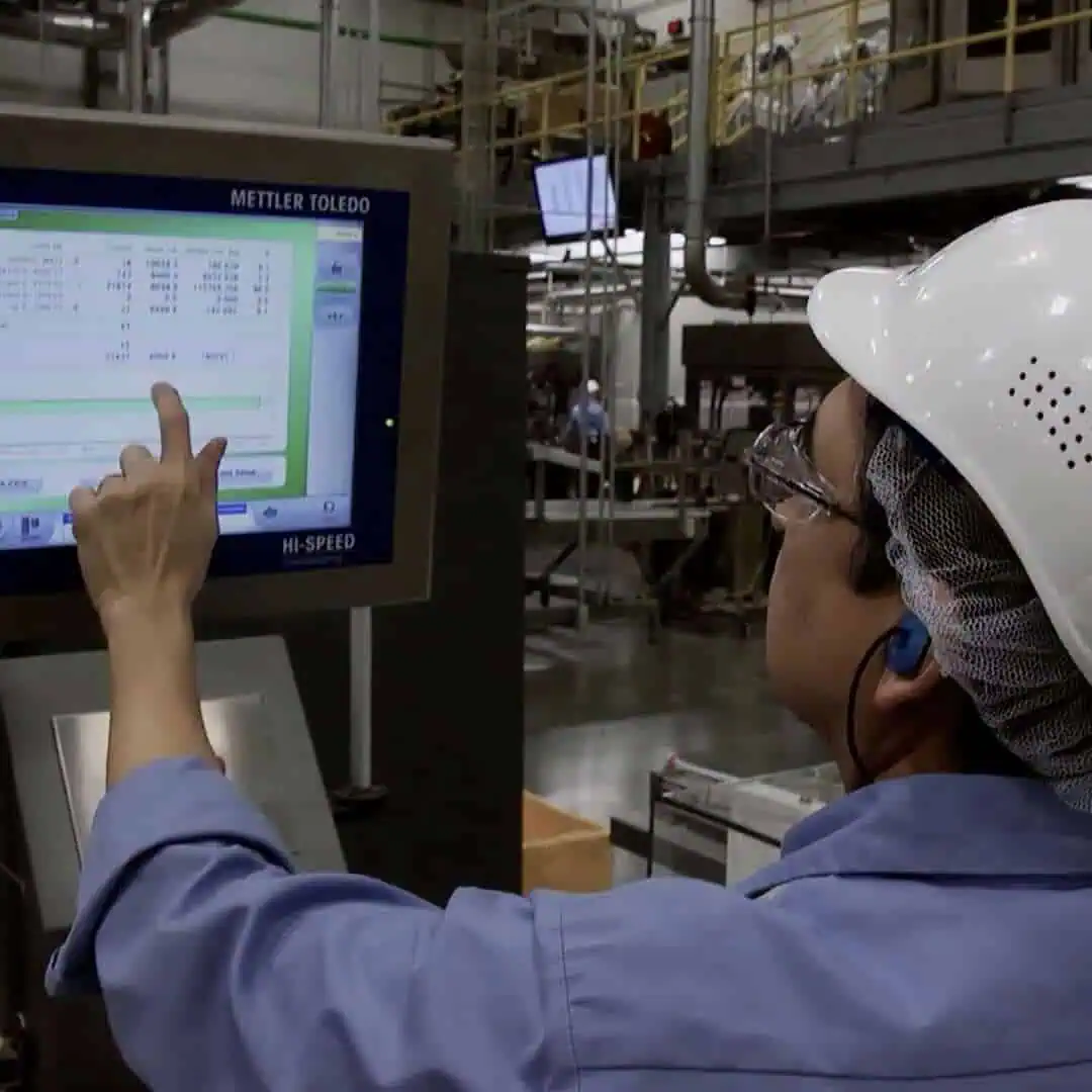 A worker in safety gear operates a touch screen on industrial equipment in a factory setting.