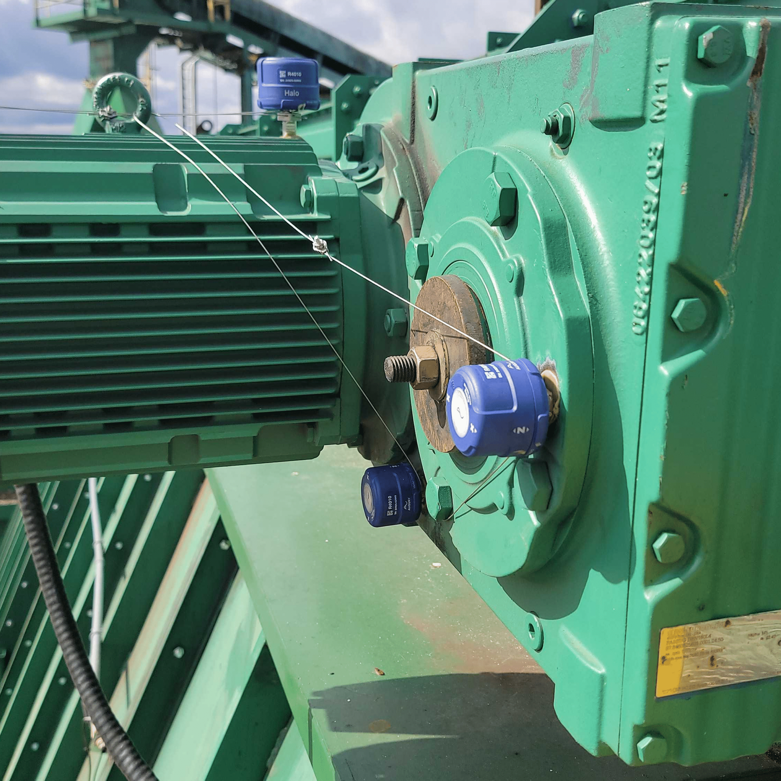 Close-up of industrial green machinery with blue devices attached and wires secured with seals, likely for monitoring or security purposes. The background shows metal structures and a partly cloudy sky.