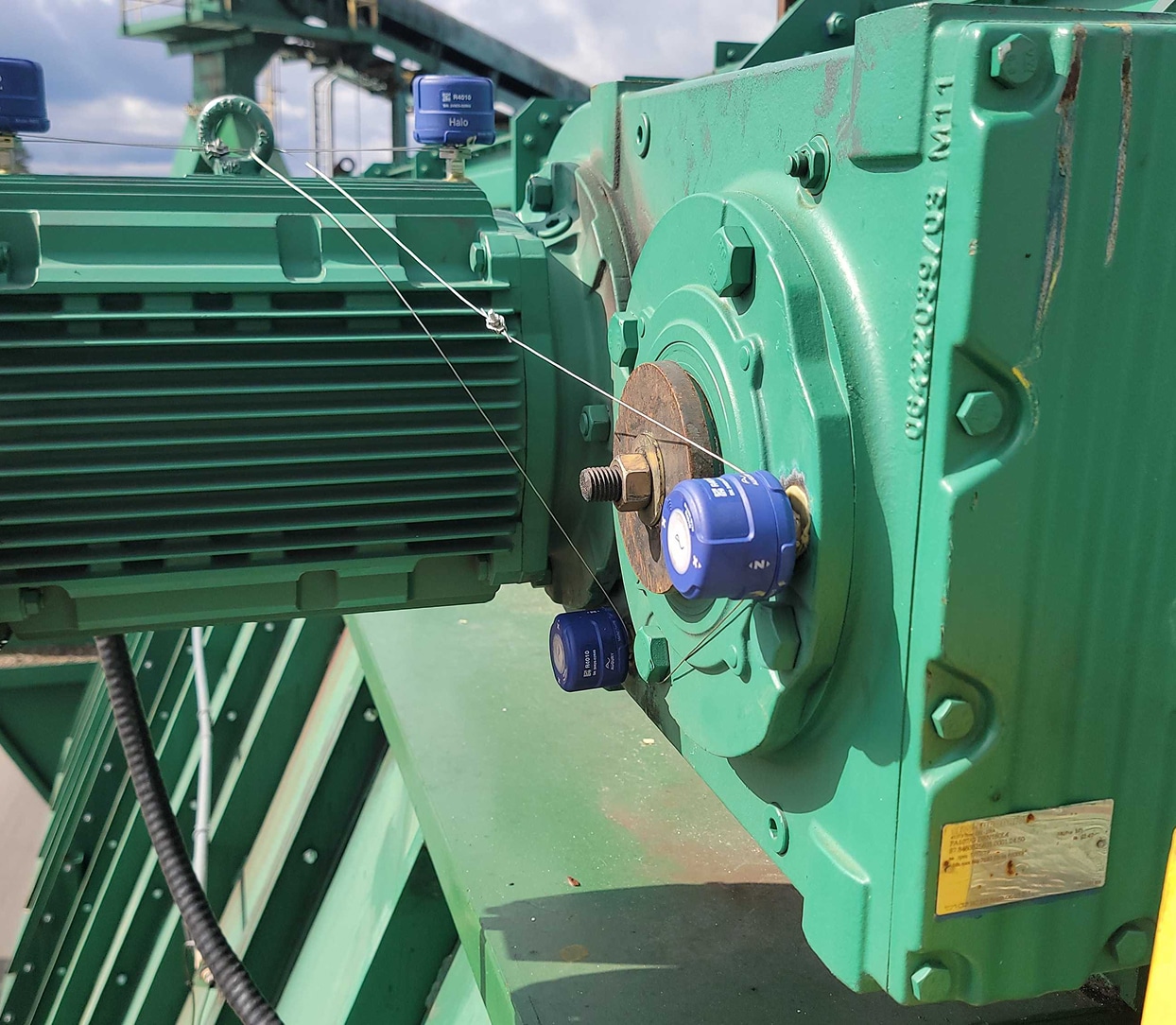 Close-up of a green industrial machine with blue monitoring sensors attached by metal wires, situated outdoors under daylight. The background shows additional green machinery and conveyor belts.