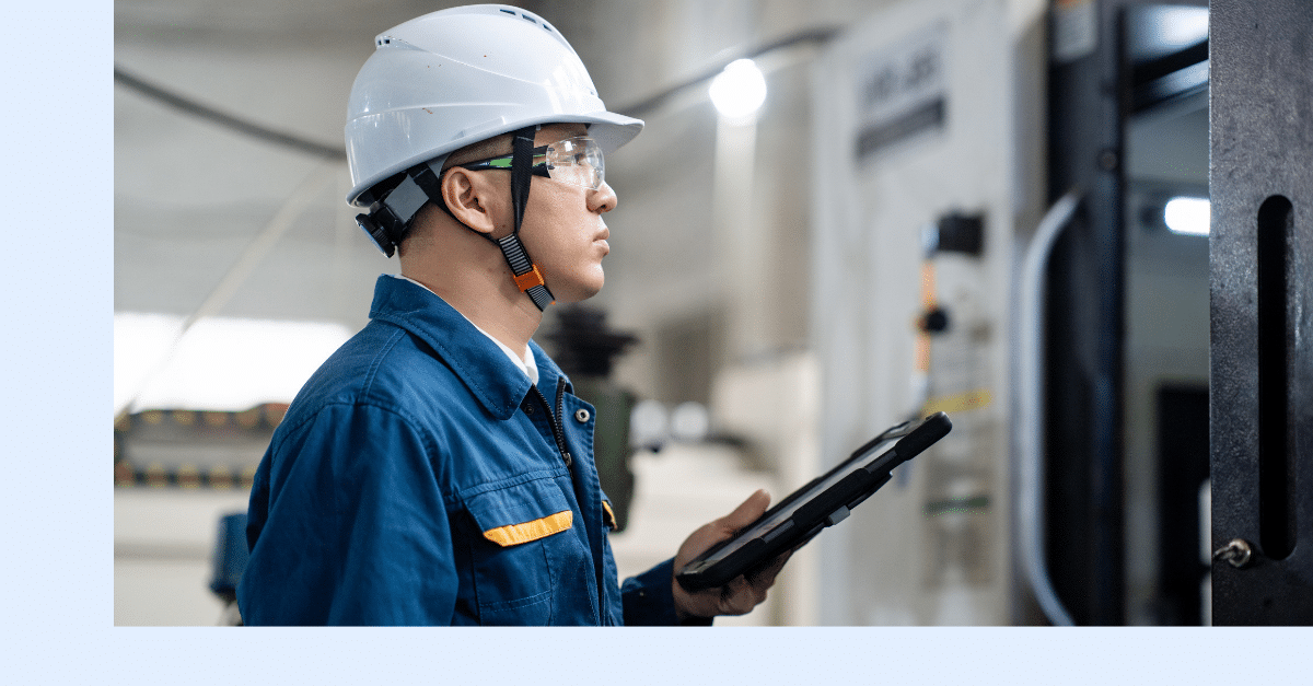 A person wearing a white hard hat, safety glasses, and a blue work uniform holds a tablet while inspecting industrial equipment in a factory setting.