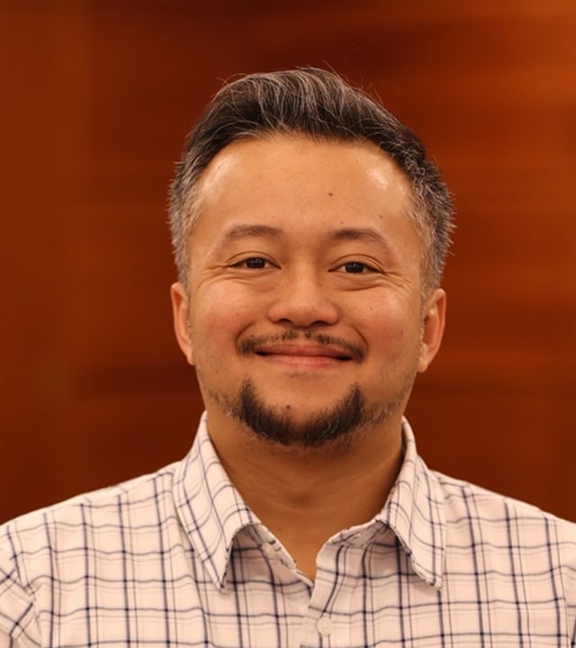 A man with short salt-and-pepper hair and a trimmed beard is smiling at the camera. He is wearing a white plaid shirt and standing in front of a blurred brown background.