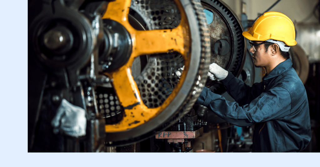 A worker in a yellow hard hat and gloves is fixing or inspecting large industrial gears inside a factory setting.