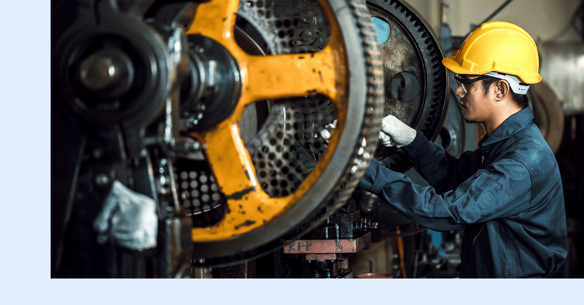A worker in a yellow hard hat and gloves is fixing or inspecting large industrial gears inside a factory setting.