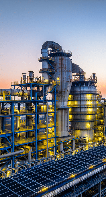 Industrial refinery plant with large metal towers, pipes, and tanks illuminated by yellow lights at sunset, against a clear sky. The structure features multiple platforms and walkways.