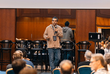 A man holding a microphone speaks on stage at a conference; several people sit in the audience, and empty chairs and reflections are visible behind him.
