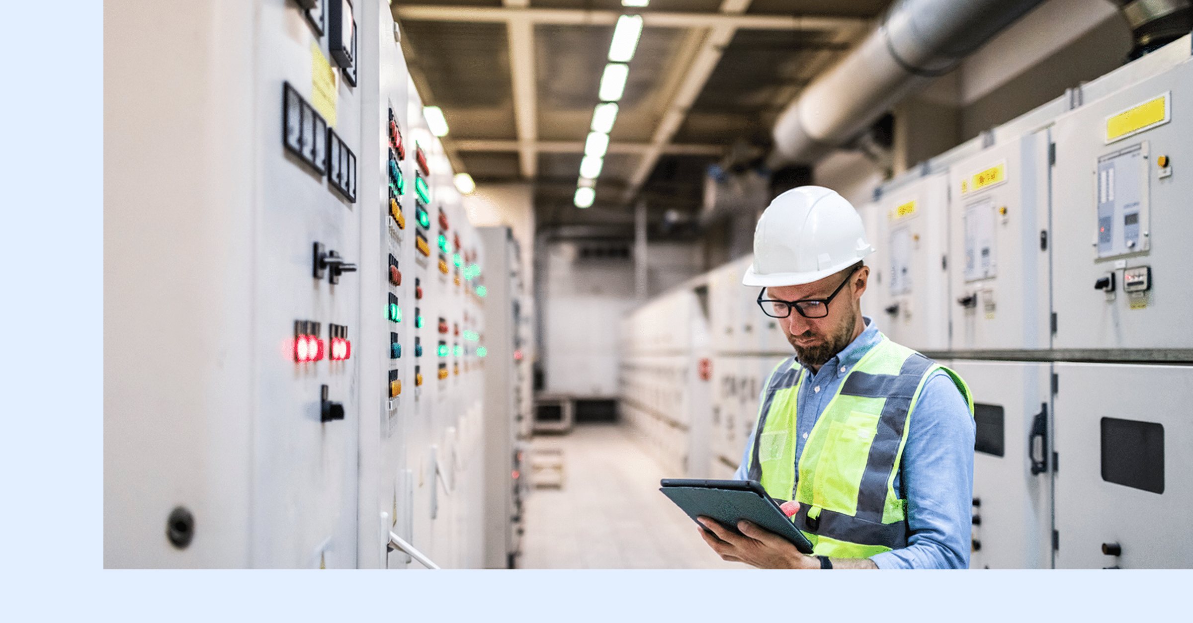 An engineer in a white hard hat and safety vest monitors a digital tablet in a control room with electrical panels.