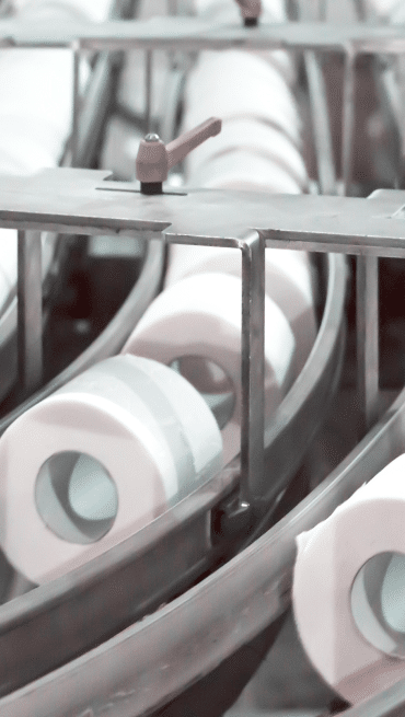 A production line from a leading tissue manufacturer showcases multiple toilet paper rolls moving along conveyor belts, with an Area Technology Manager examining the processes and a valve handle visible in the background.